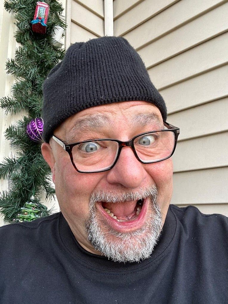 A man wearing glasses and a beanie is making a funny face in front of a christmas tree.
