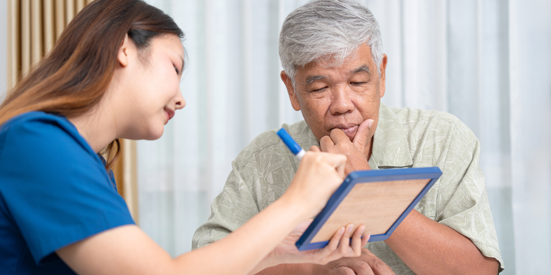 Woman in blue scrubs writes on a clipboard as an older man in a light green shirt looks on.