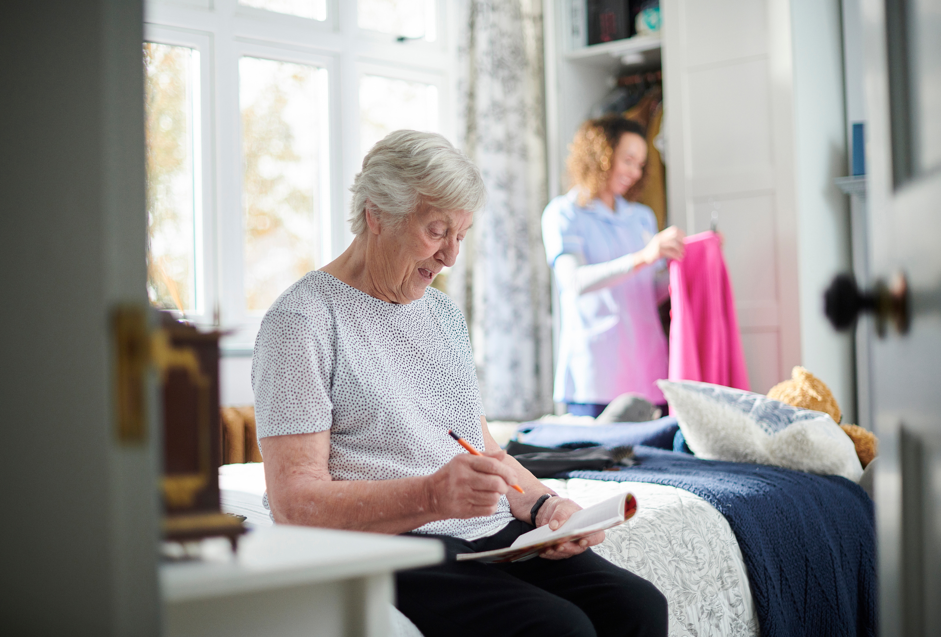 Elderly person sitting, writing on a notepad. Caregiver in background folds clothes in a bright room.