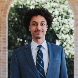 Man in blue suit and tie stands in front of brick archway. He smiles.