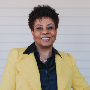 Woman in yellow blazer smiles, stands outdoors in front of white siding.