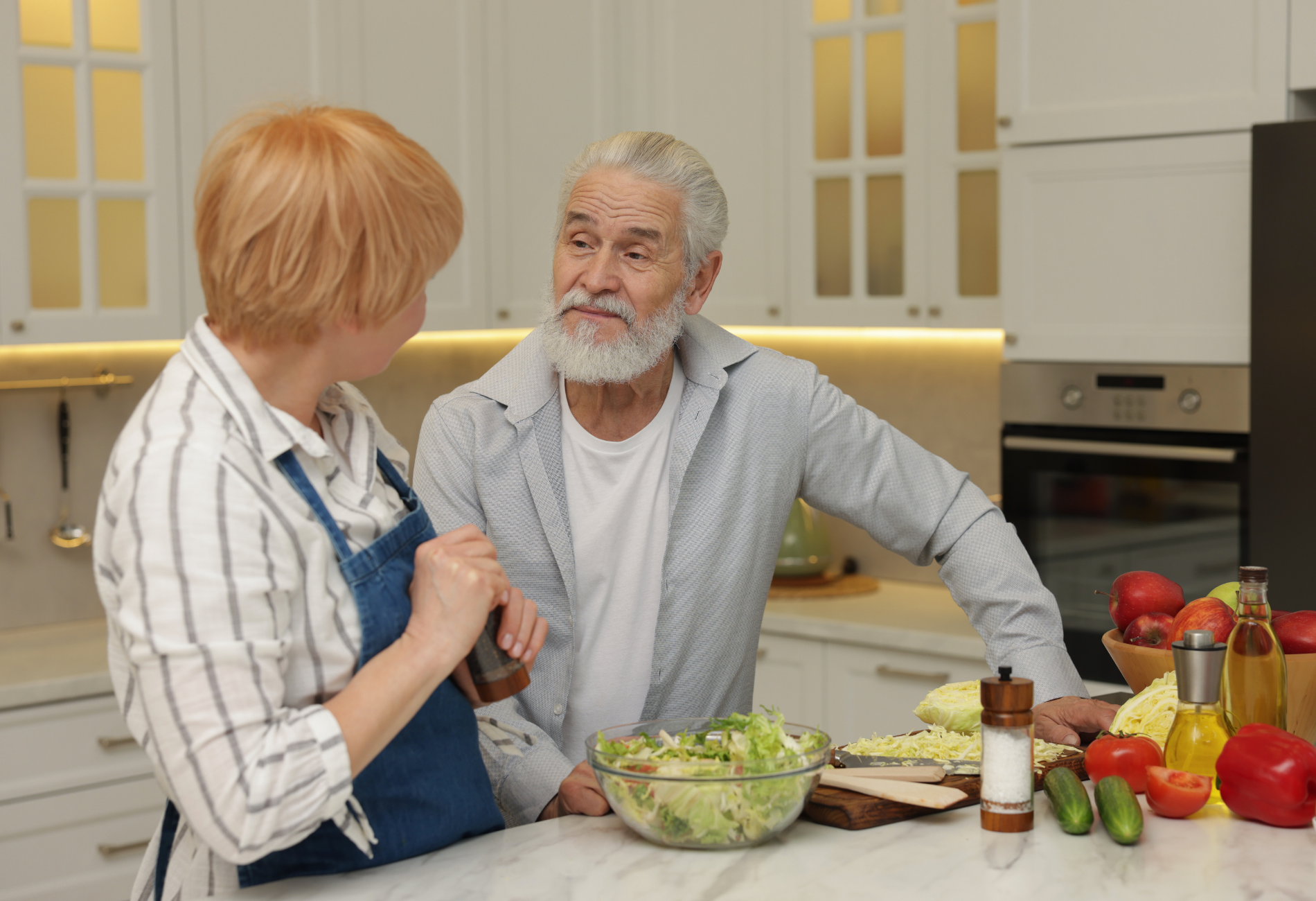 Couple in kitchen preparing food, chatting near a salad bowl and vegetables.