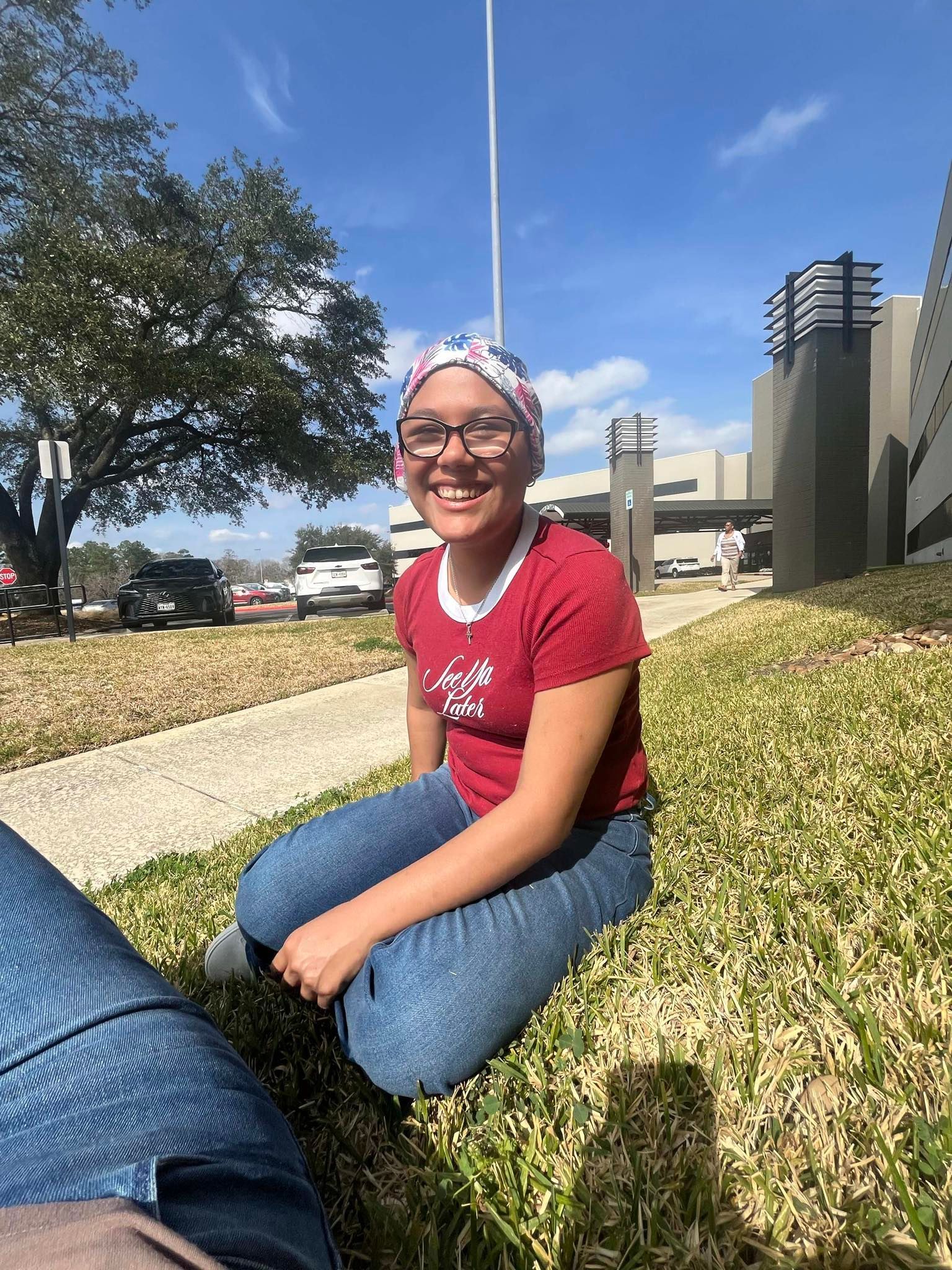 A smiling person in a red shirt and floral head covering sits on a grassy lawn in front of a building on a sunny day.