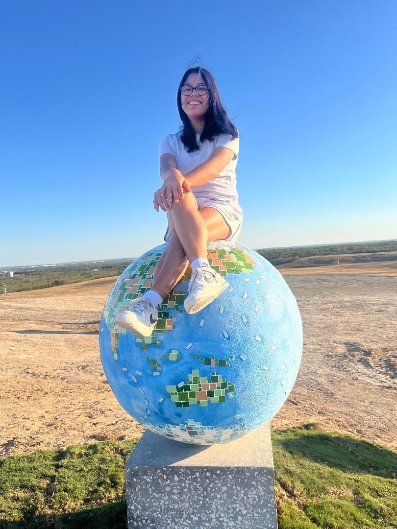 A person sits smiling atop a globe sculpture in an outdoor, sunny setting.
