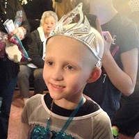 A smiling child wearing a silver toy crown and a teal garment, with other people blurred in the background.