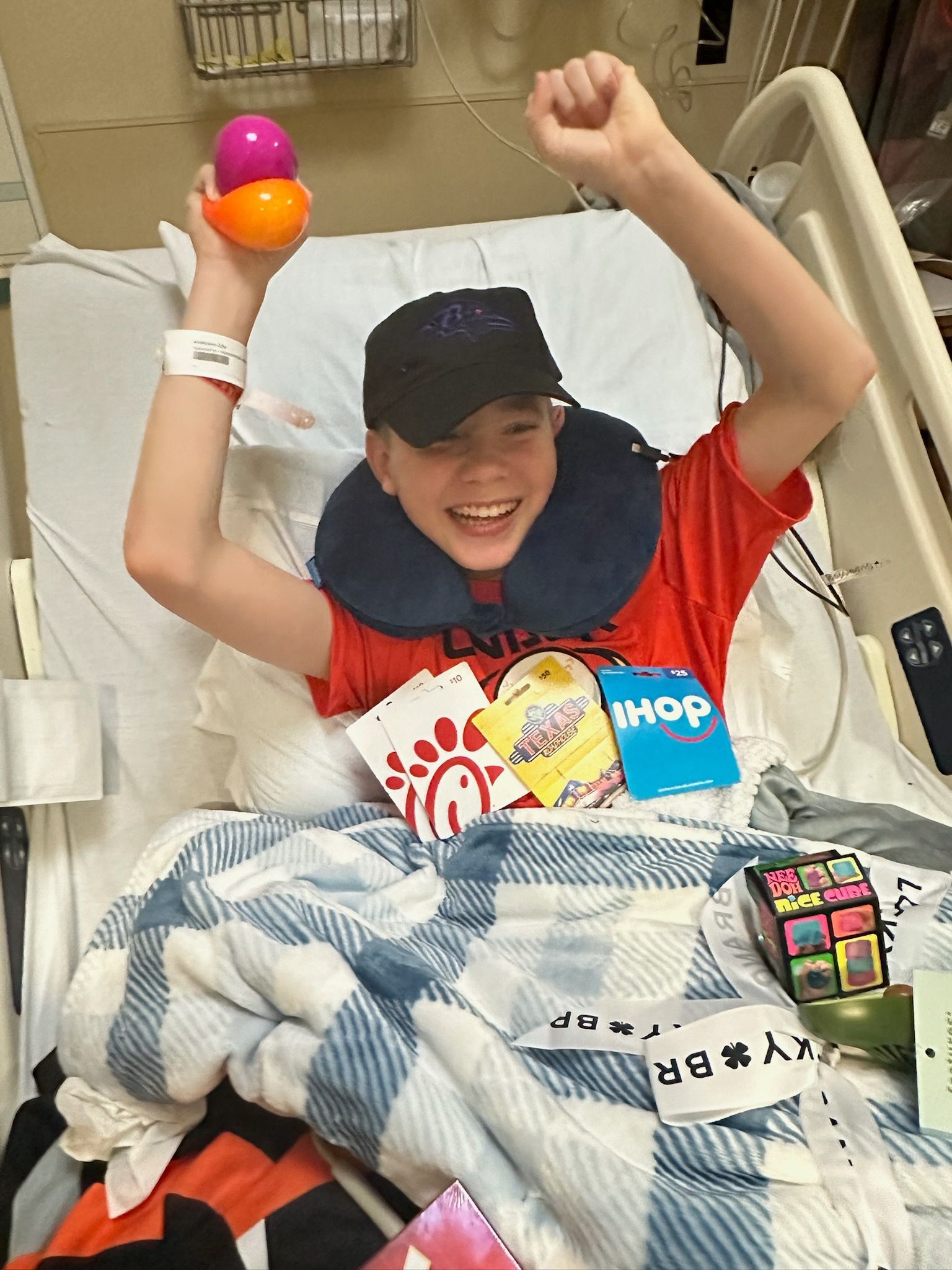 A smiling person sits in a hospital bed, holding plastic eggs, wearing a red shirt and hat with food gift cards nearby.