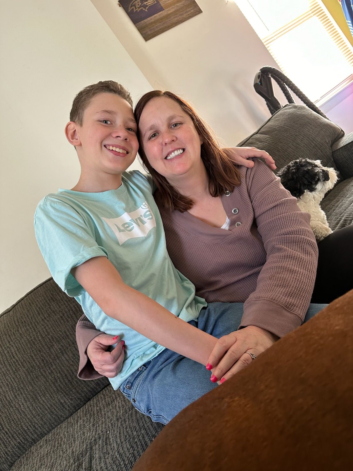 A person in a light blue t-shirt and another in a mauve sweater embrace on a couch next to a small dog.