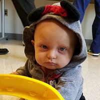 A baby wears a grey Minnie Mouse hoodie with ears and a red bow, looking directly forward, with a yellow toy in the foreground.