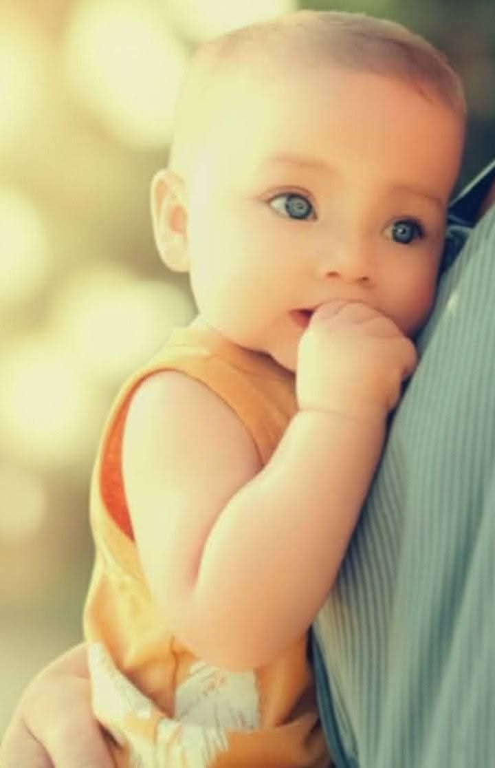 A baby in a mustard-colored top resting on a person’s shoulder, looking to the side with a hand near their mouth.