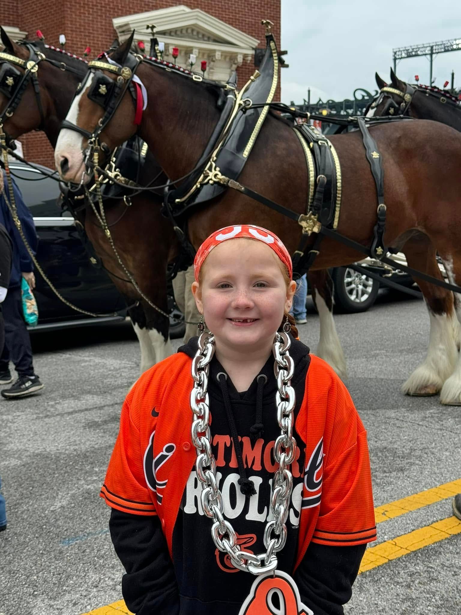 A child wearing an Orioles jersey and chain necklace stands smiling in front of a pair of draft horses in harnesses.