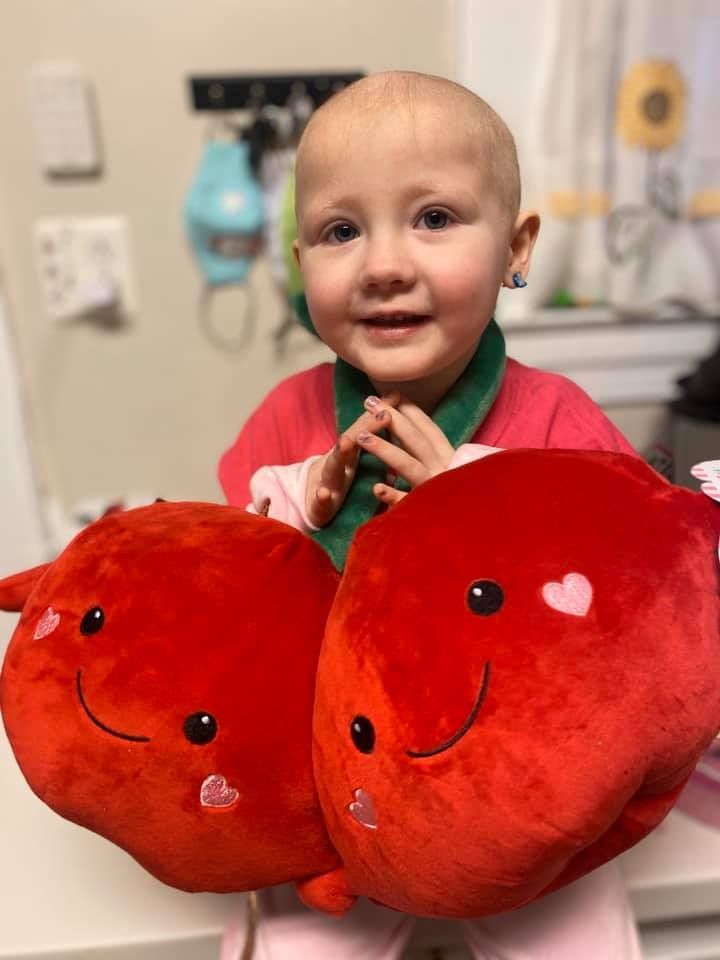 A smiling child holds two large, bright red, heart-shaped plush toys, each decorated with small pink heart accents.