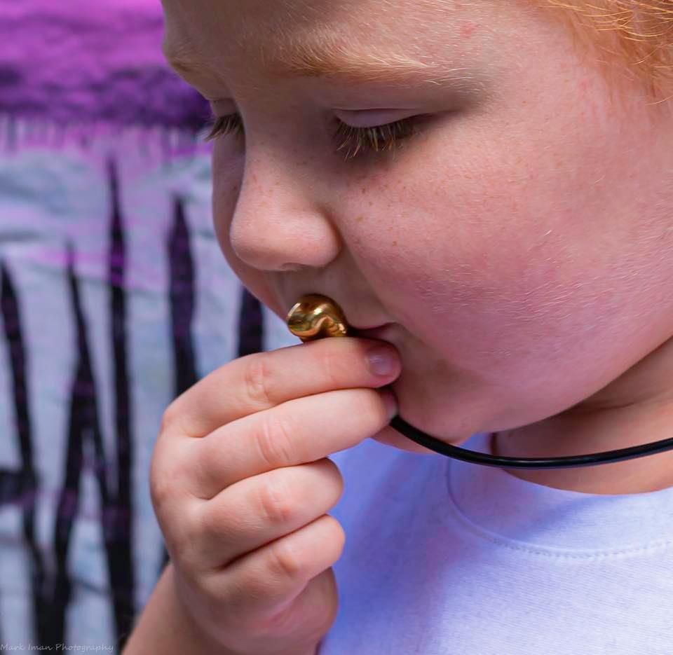 A person with light hair holds a small, gold, shell-shaped pendant against their lips, wearing it on a black cord.