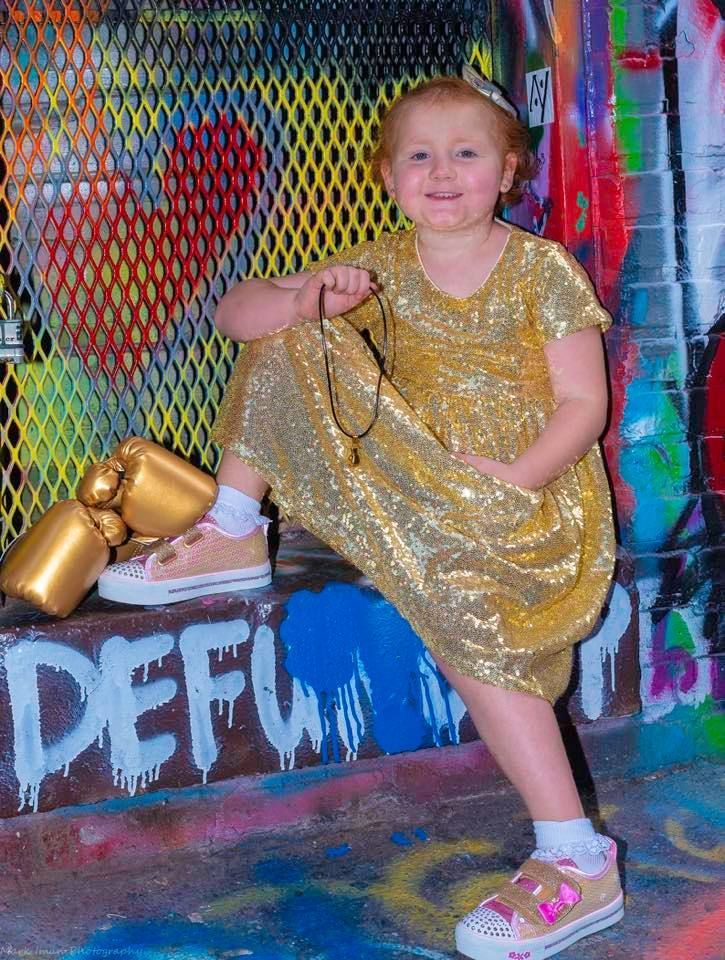 A person in a gold sequined dress sits on a concrete ledge with graffiti, holding a small gold object and wearing sneakers.