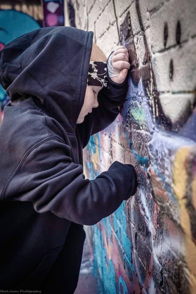 A person in a black hooded sweatshirt with a patterned headband stands leaning against a colorful graffiti brick wall.