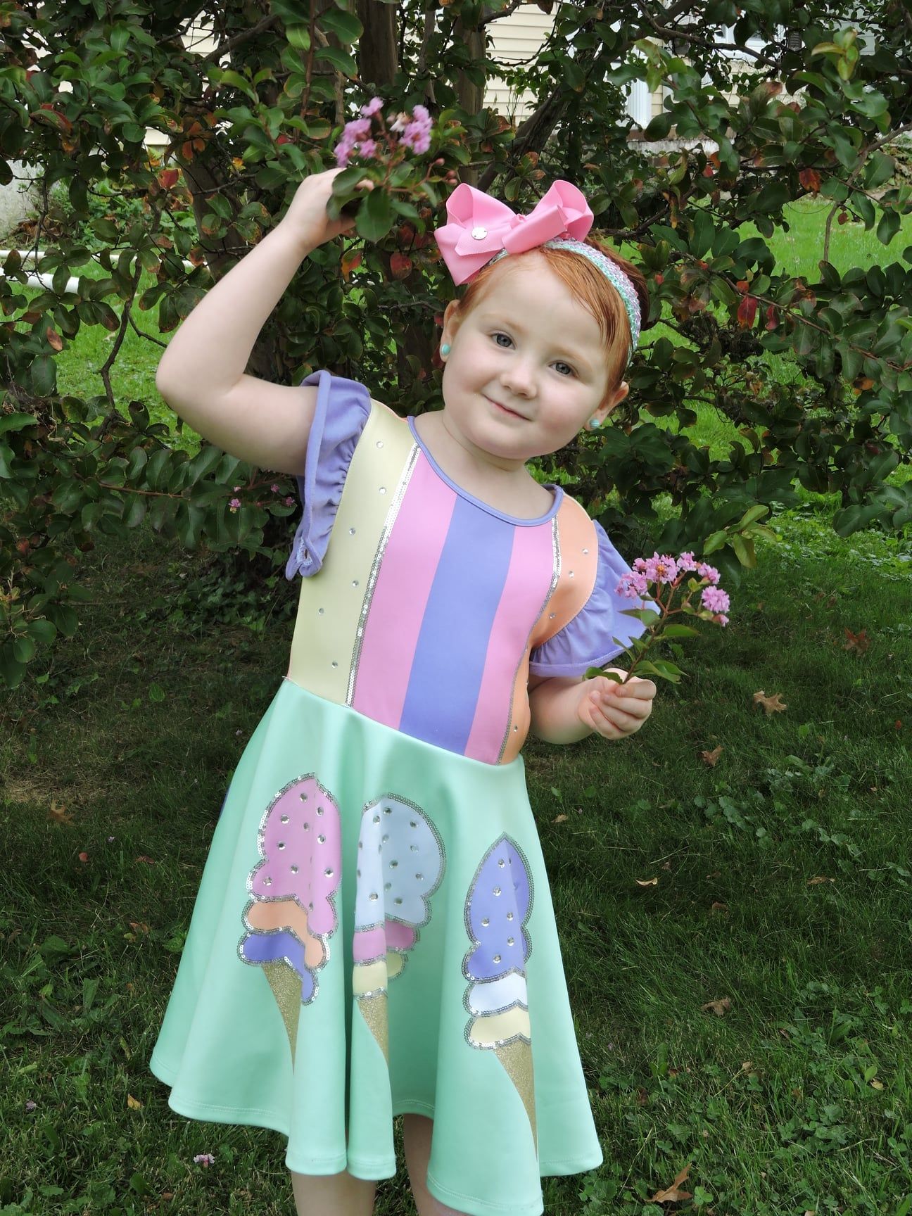 A smiling child in a pastel ice-cream-patterned dress and a large pink hair bow, standing outside near a leafy tree.