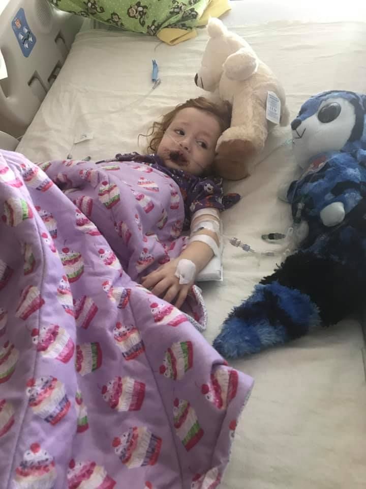 A child lying in a hospital bed with an IV, wearing a cupcake-print blanket and resting beside two stuffed animals.