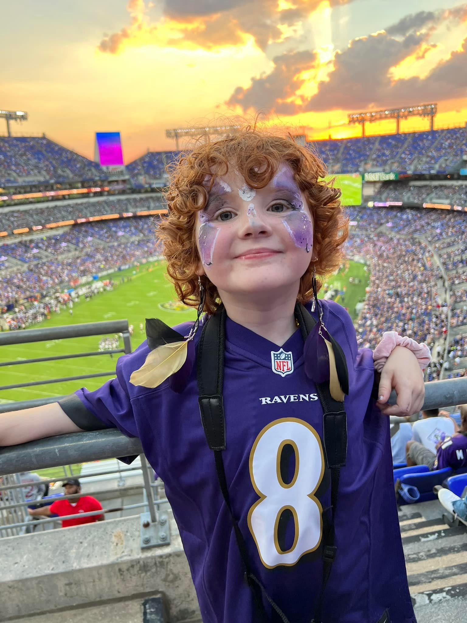 A person with curly hair and face paint wears a purple number 8 Baltimore Ravens jersey at a sunny stadium.