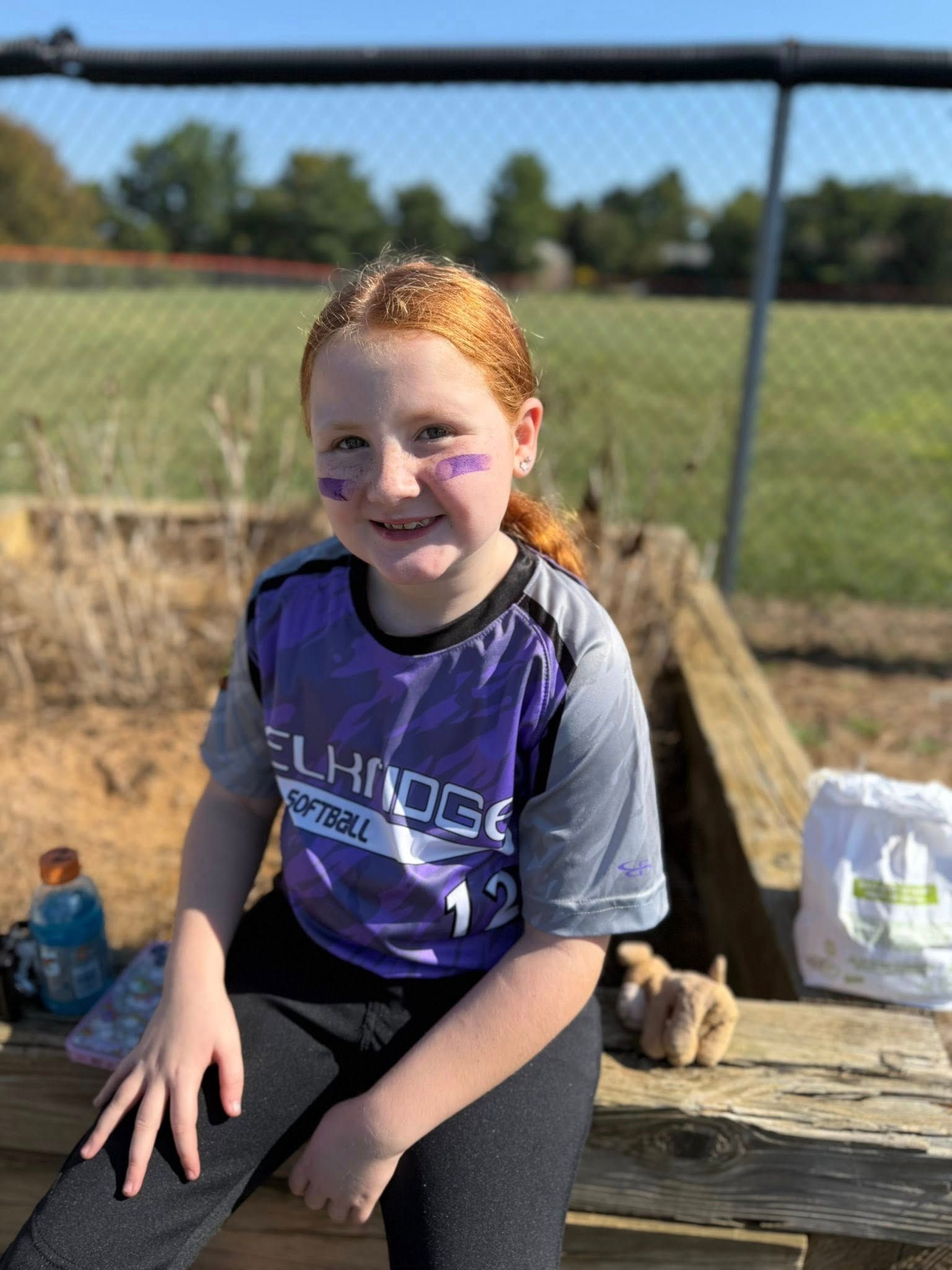 A softball player wearing a purple jersey with eye black sits on a wooden bench at a field on a sunny day.