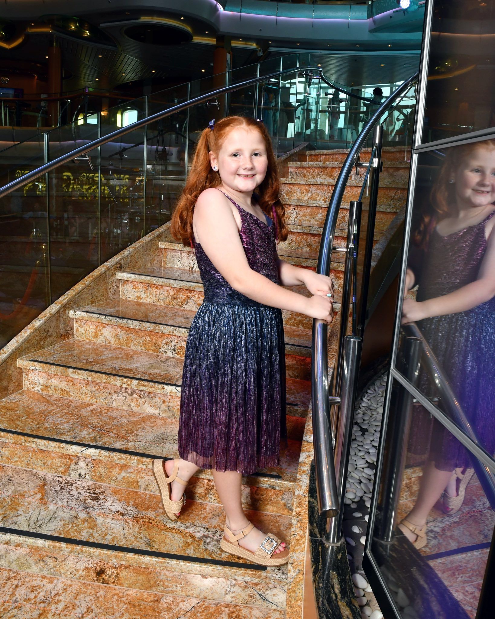 A smiling child with red hair in a purple dress stands on a marble staircase, holding onto a silver railing.