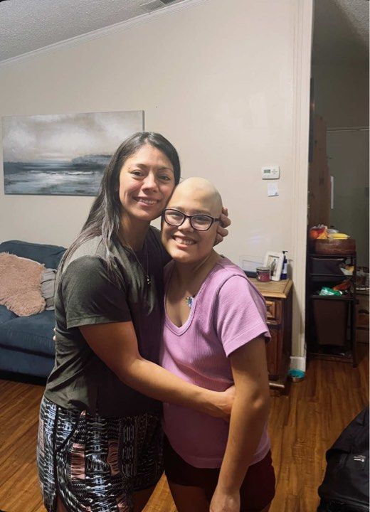 Two people embrace and smile in a living room, one wearing a dark shirt and the other in a light purple shirt with a shaved head.