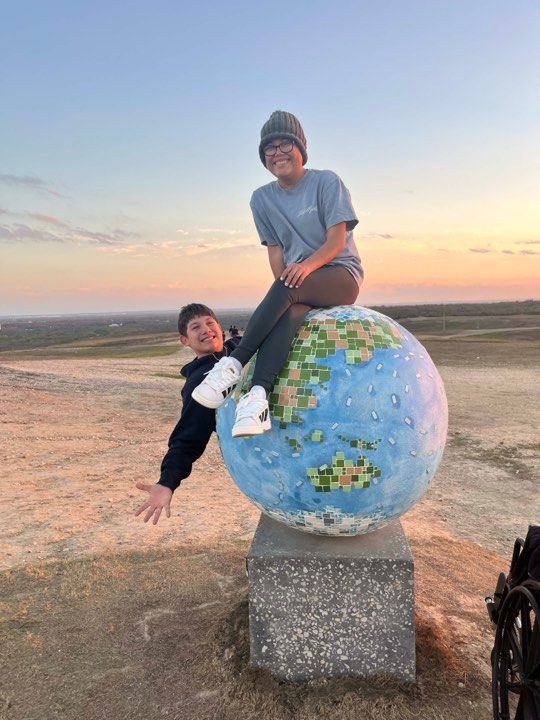 Two people pose by a globe sculpture on a pedestal at sunset, one sitting on top and the other waving from behind it.