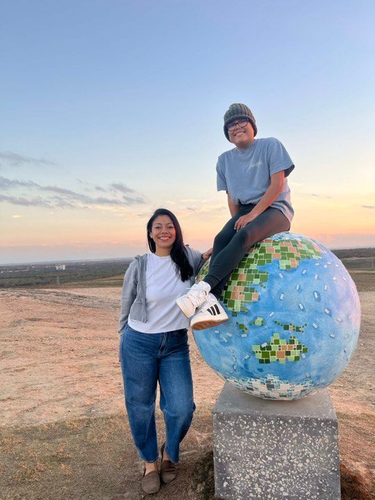 A person standing next to another person sitting on top of a large, painted globe sculpture at sunset.