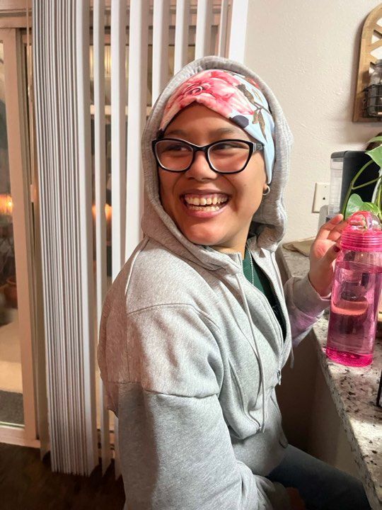 A smiling person wearing a grey hoodie and a floral head covering, holding a pink water bottle in a kitchen setting.