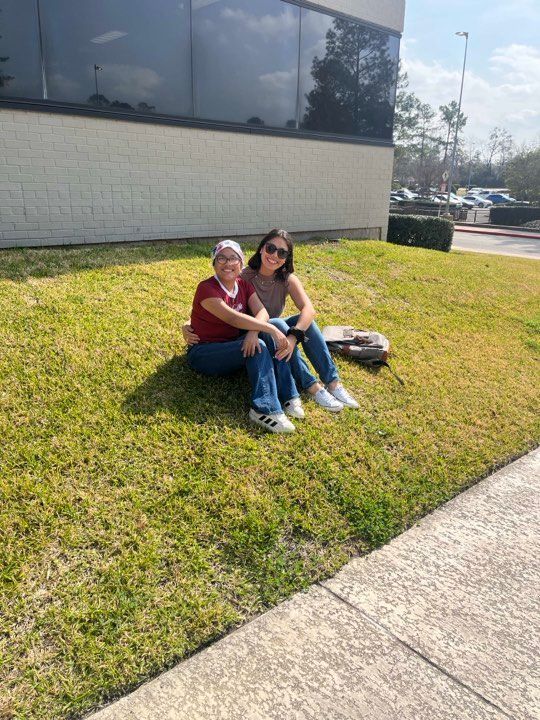Two people sit smiling together on a grassy lawn next to a building on a sunny day.