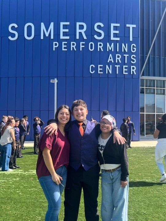 Three people smile while posing in front of the blue Somerset Performing Arts Center building on a sunny day.