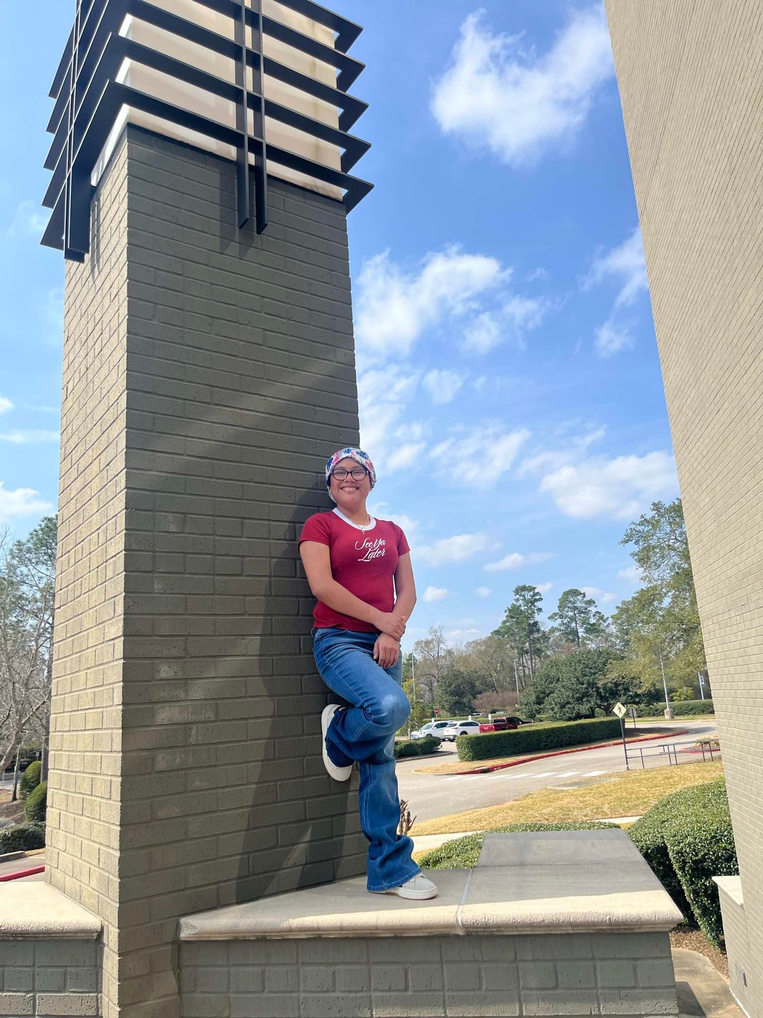 A person wearing a red shirt and jeans leans against a tall, grey brick column outdoors under a bright blue sky.