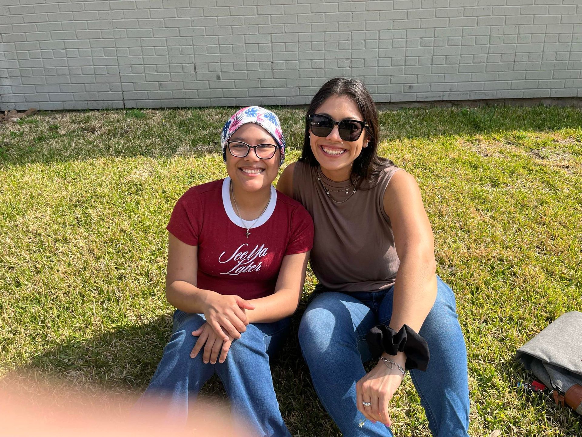 Two people sit smiling on a grassy lawn in front of a gray brick wall on a sunny day.