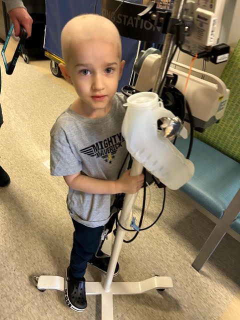 A child stands in a medical setting, holding a medical IV pole for support.