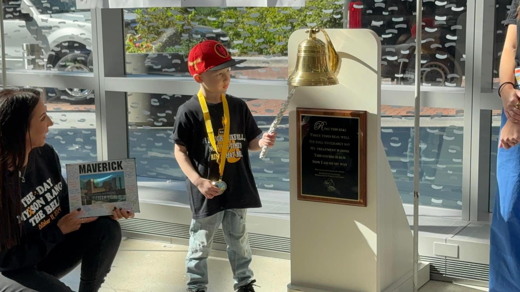 A child in a red cap and yellow lanyard rings a brass bell next to a sign, accompanied by an adult holding a photo.