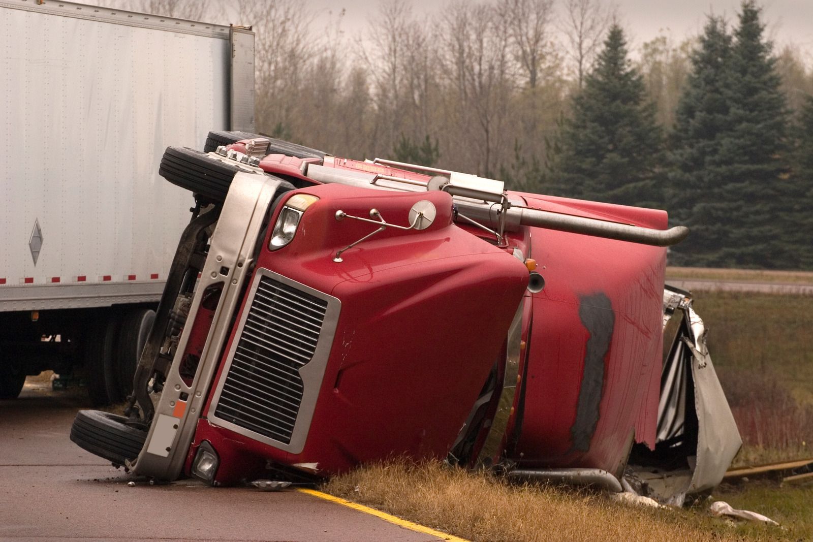Red semi-truck overturned on its side by a road, trailer to the left.