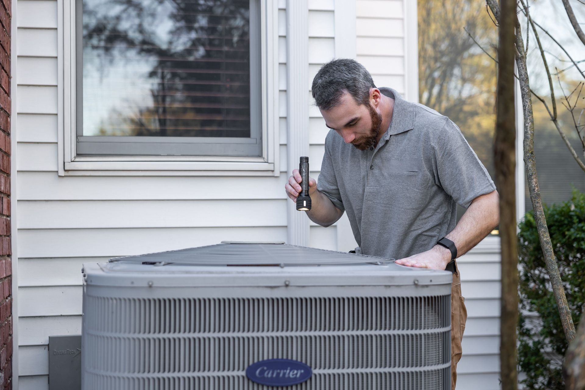 Technician inspecting an outdoor Carrier HVAC unit beside a house window