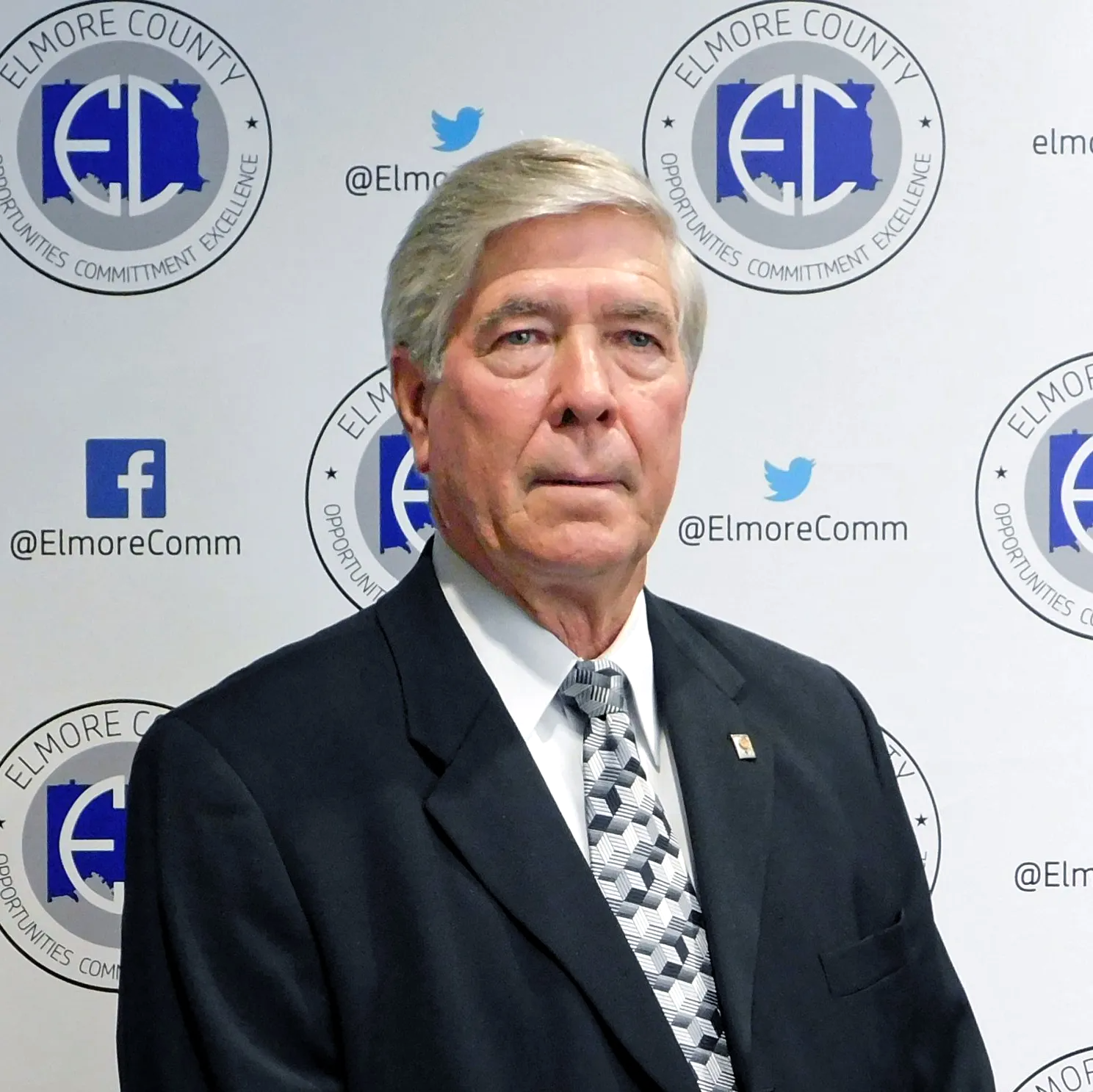 Man in a suit, likely a public official, poses in front of a backdrop with the Elmore County logo and social media handles.