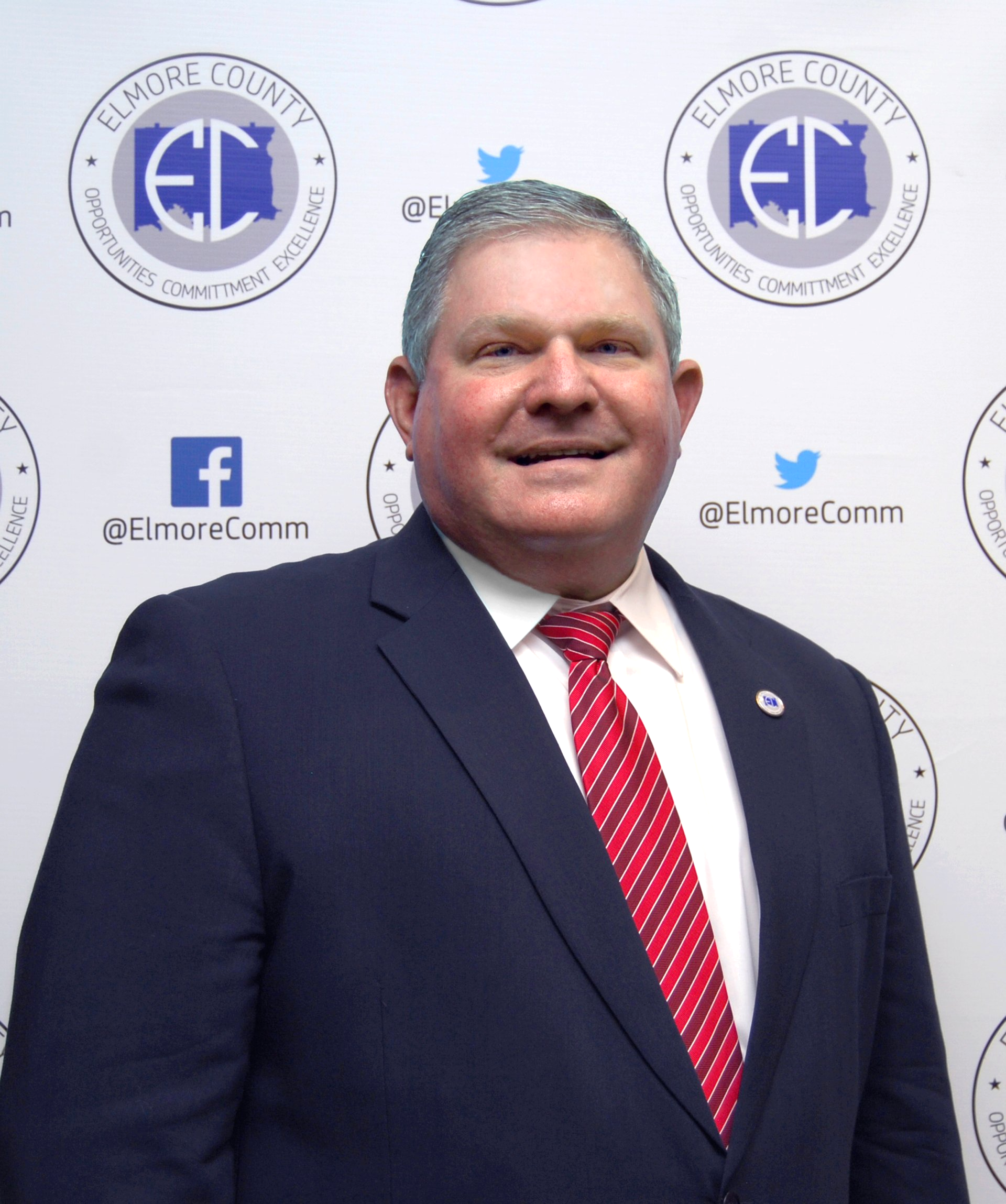 Man in a navy suit and red tie smiles at the camera, standing in front of a wall with Elmore County logos.
