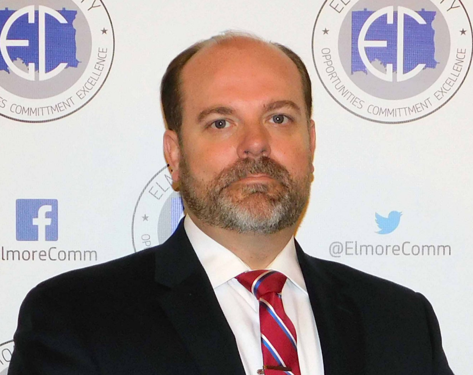 A middle-aged man with a beard, wearing a suit and tie, stands in front of a backdrop with the Elmore County logo.