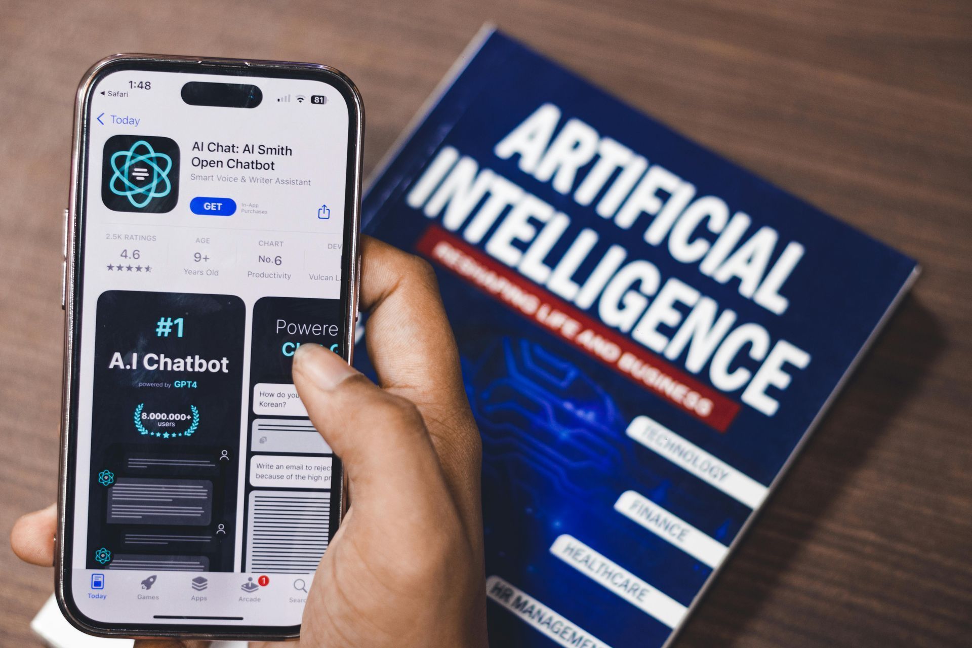 Hand holding a smartphone beside a book titled “ARTIFICIAL INTELLIGENCE” on a wooden table.