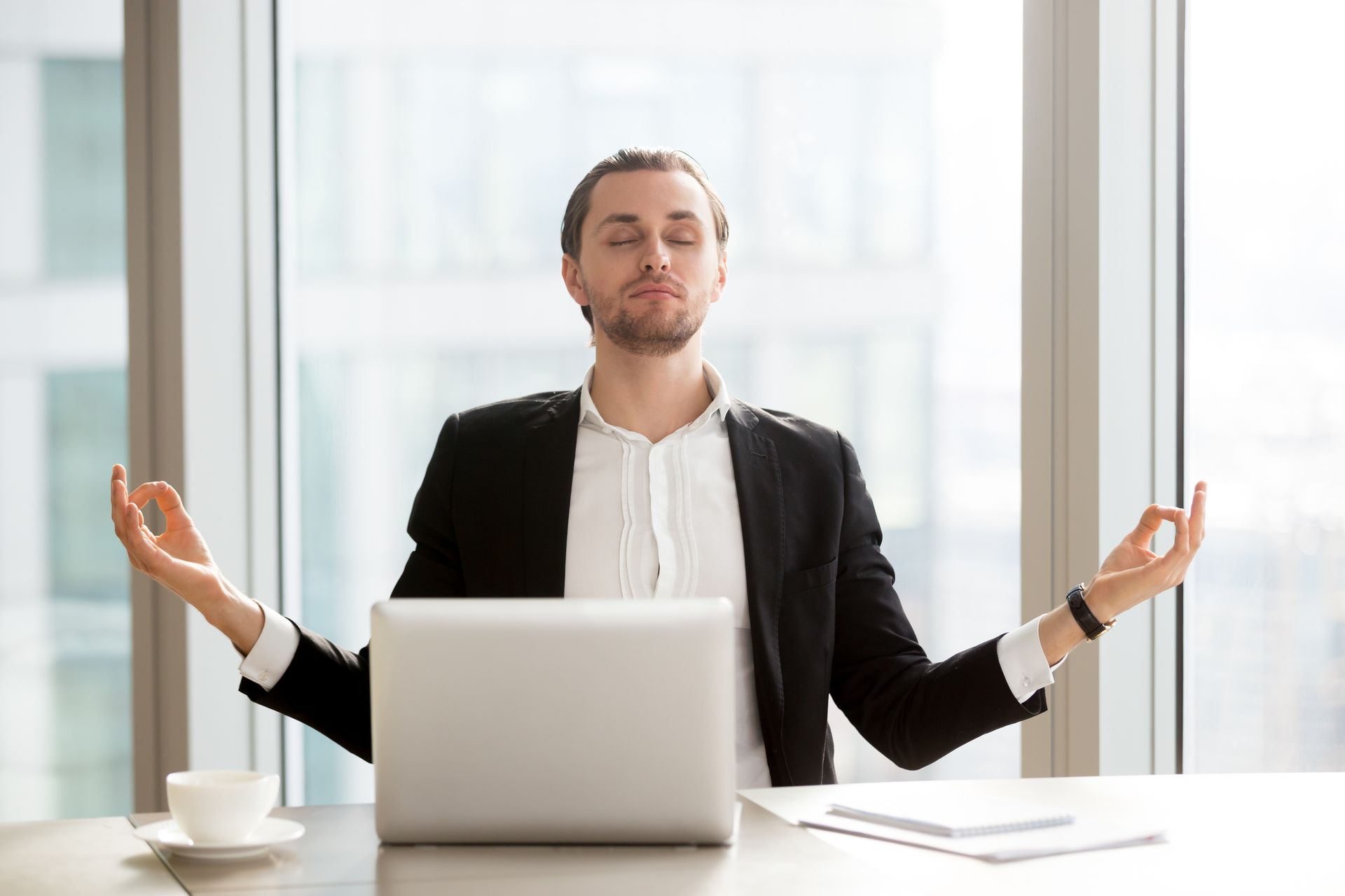 A man is meditating at a desk in front of a laptop computer.