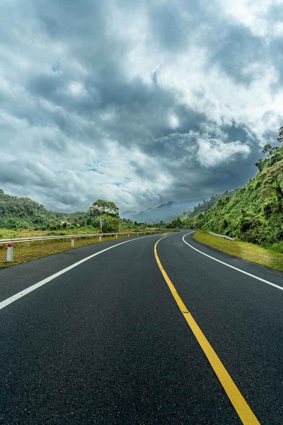 Curving empty highway under dramatic cloudy sky, with lush green roadside vegetation.