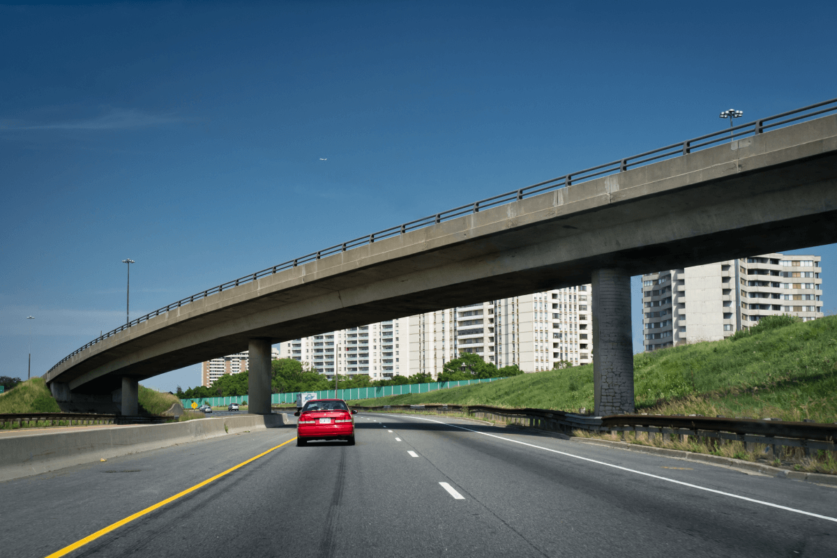 Red car driving on a highway beneath a concrete overpass near apartment buildings