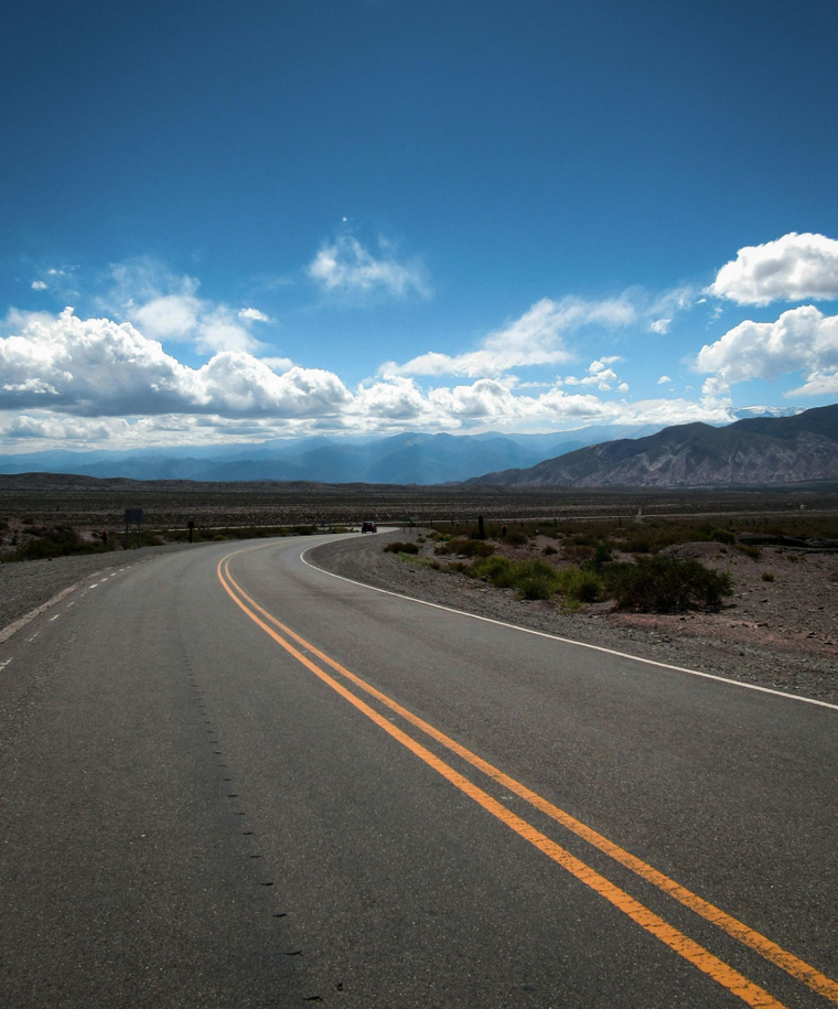 Curving empty highway through desert landscape under a blue sky with clouds and distant mountains