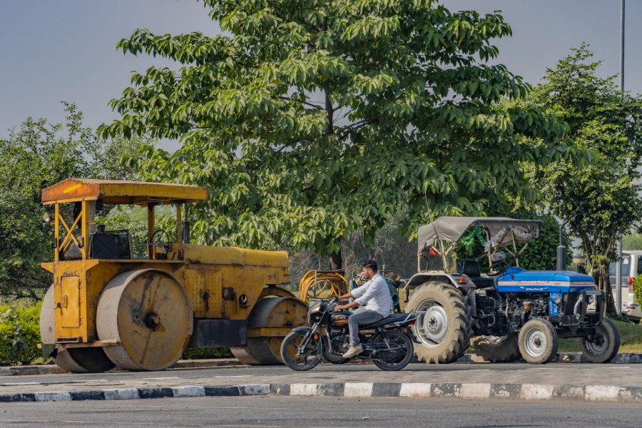 Yellow road roller and blue tractor stopped by a roadside, with a motorbike rider nearby under trees.