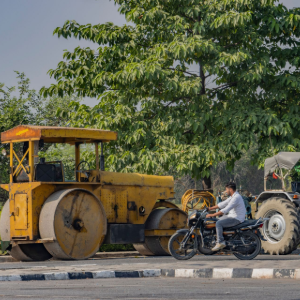 Yellow road roller parked by a man on a motorcycle near a tractor on a roadside