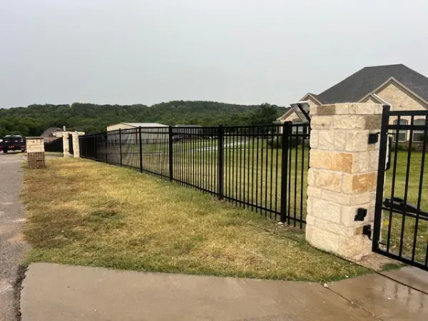 A black metal fence surrounds a brick pillar in front of a house.