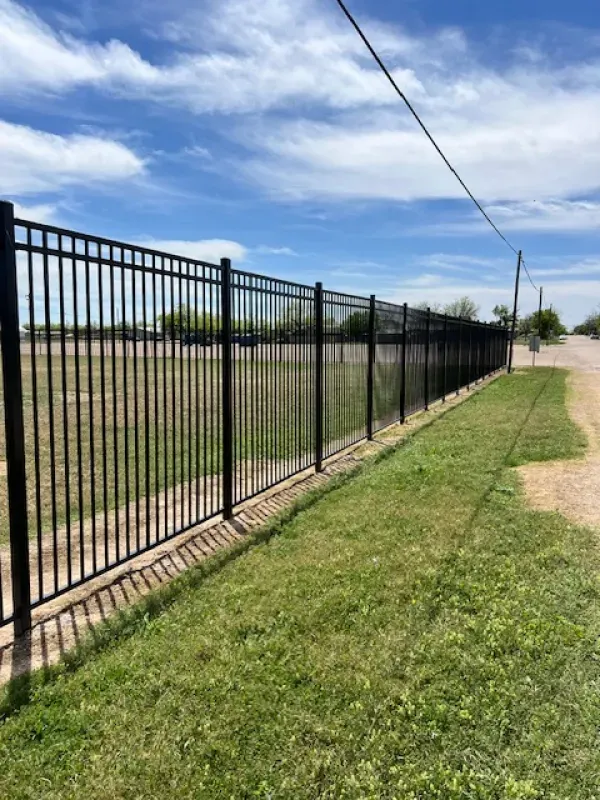 A black metal fence surrounds a grassy field next to a dirt road.