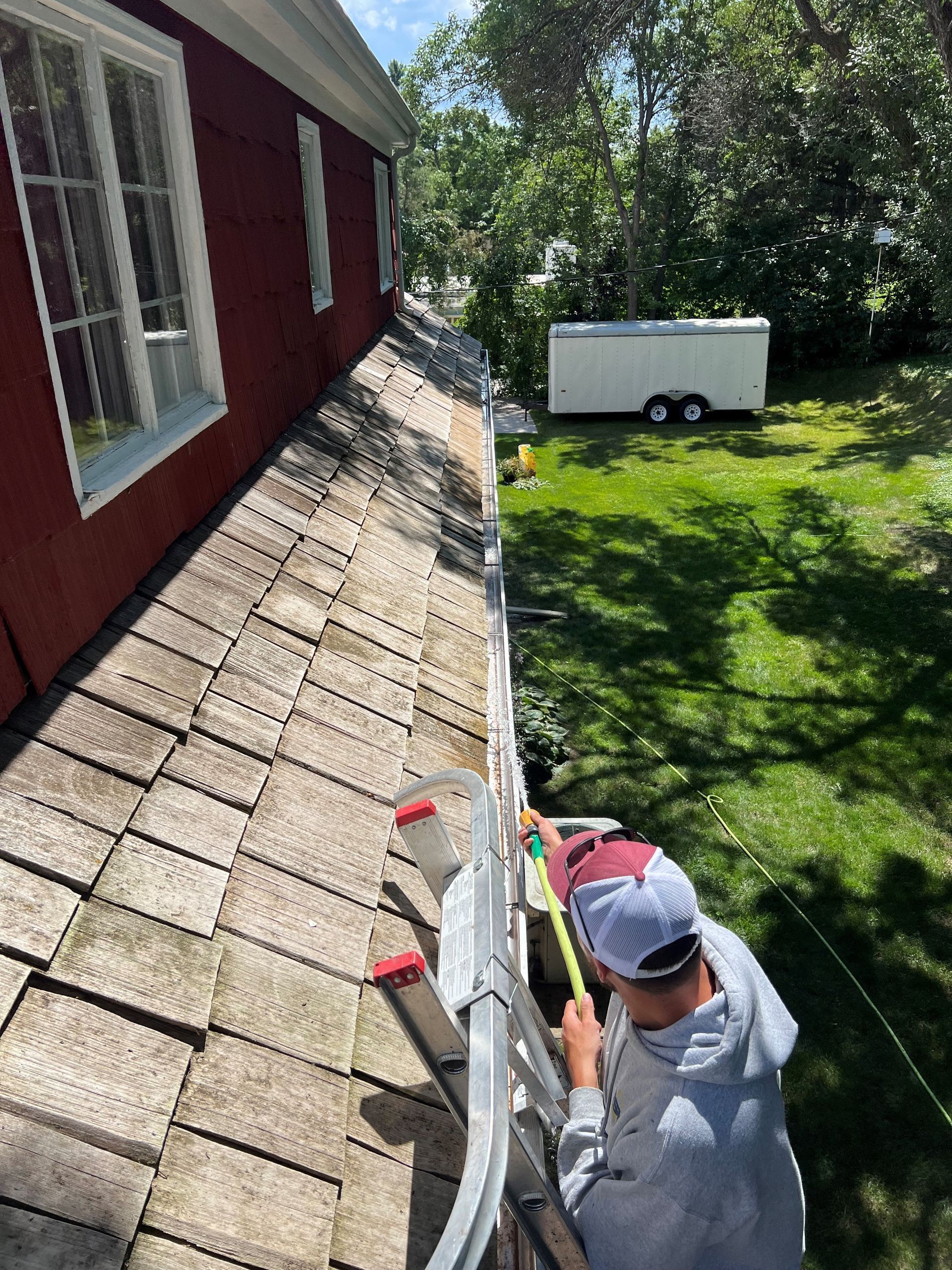 Person on a ladder cleaning a gutter on a red house with a green lawn and a trailer in the background.