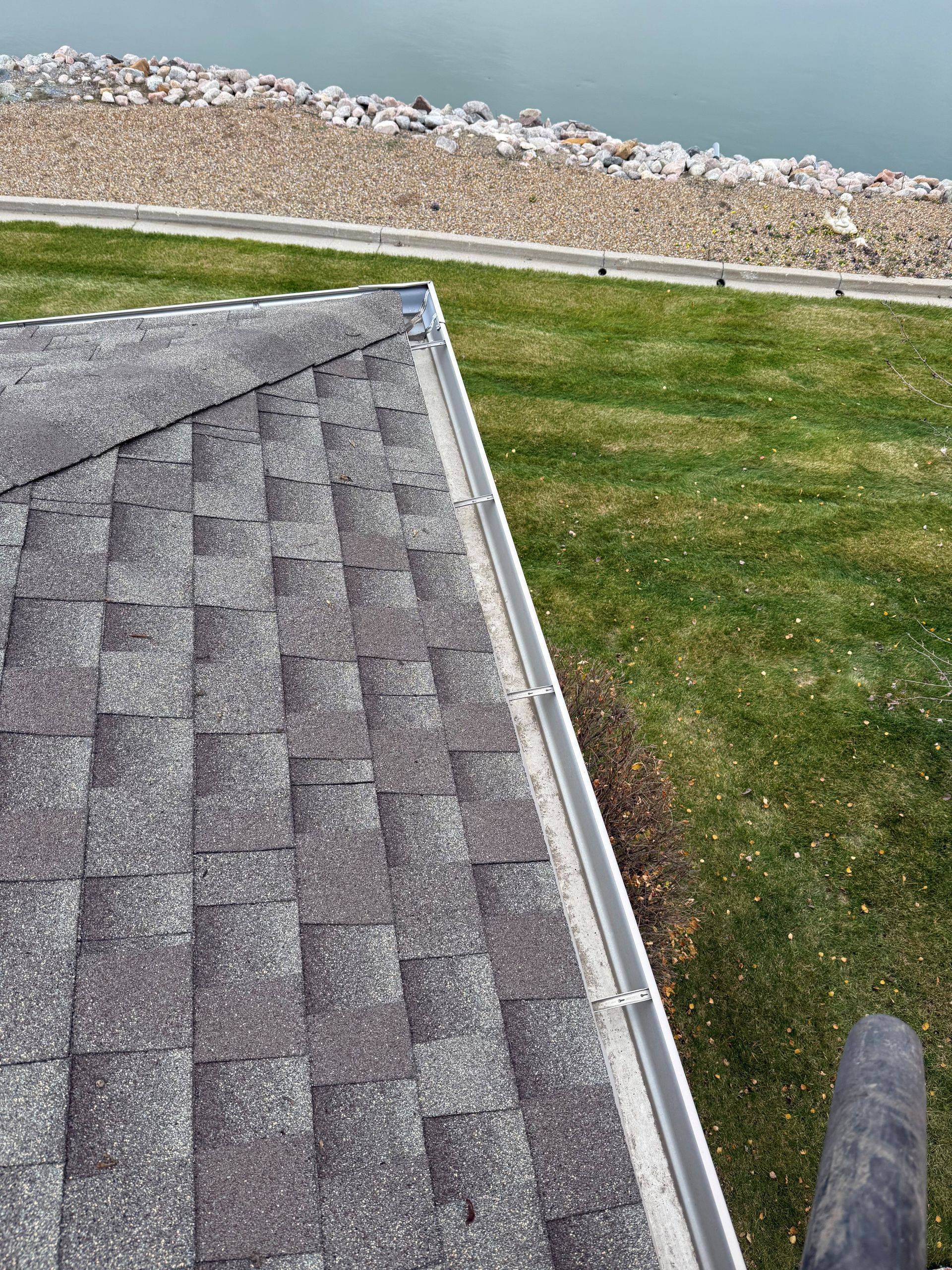 A roof with gray shingles and a gutter, overlooking grass and a water body with rocks in the background.