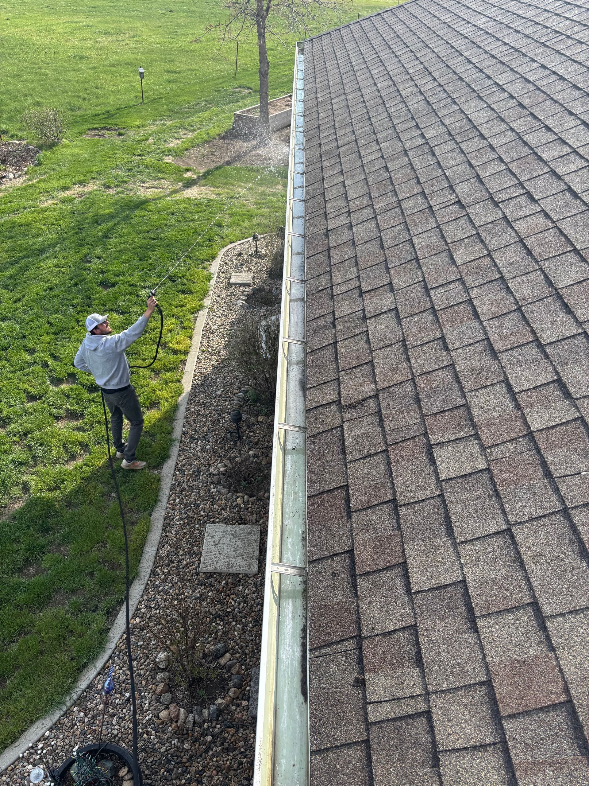 Man washing roof gutters with a spray nozzle. Green grass, gravel, and a roof are also visible.